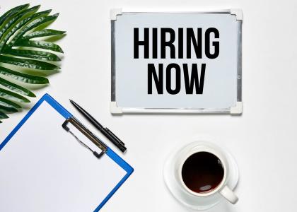 Hiring Now sign next to plant, coffee cup and clipboard